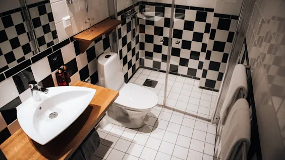 Håndvask med en træbordplade er placeret ved en toiletkumme; brusebad med glasdøre er ved siden af. Sorte og hvide fliser pryder badeværelsesvægge og -gulv.