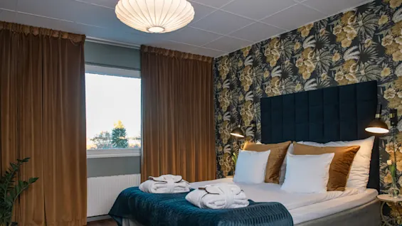 A neatly made bed with folded robes sits in a room with floral wallpaper, brown curtains, and a window overlooking trees, lit by a ceiling lamp.