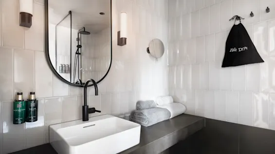 Ett modernt badrum har ett vitt handfat med svart kran, under en oval spegel. Handdukar och en "Hair Dryer"-påse ligger på en mörk bänkskiva. Minimalistisk kaklad bakgrund.