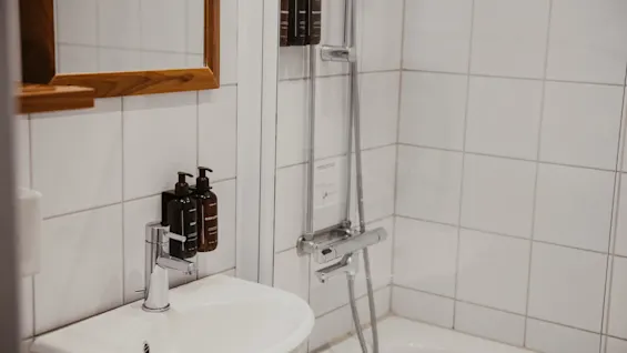Et rent badeværelse med en hvid vask, spejl og flisebelagt bruseområde. Body wash og shampoo dispensere er tilgængelige.