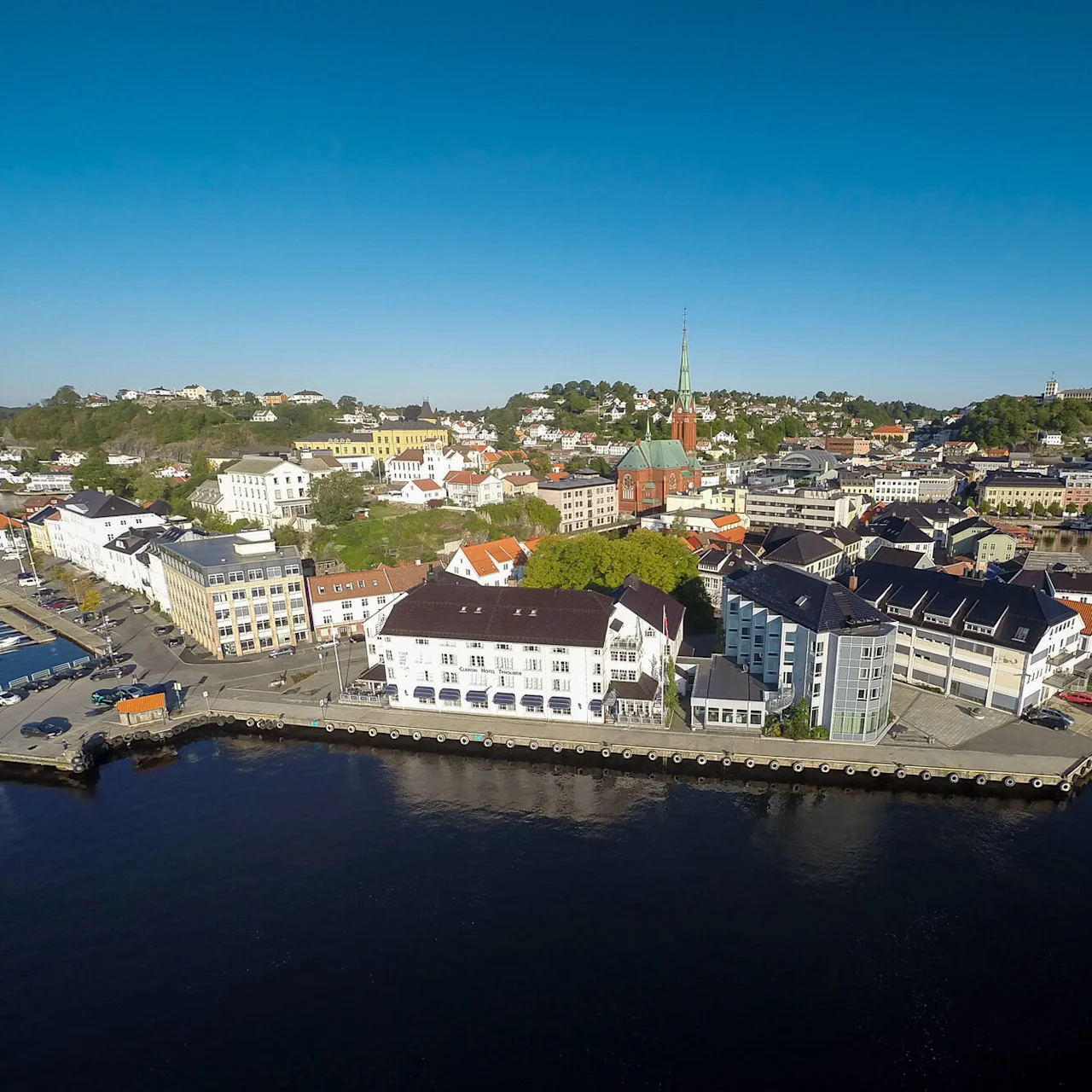 Bygningene i kystbyen har utsikt over en rolig havn med flere båter fortøyd; et høyt kirkespir reiser seg blant grønne åser under en klar blå himmel.