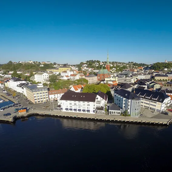Bygningene i kystbyen har utsikt over en rolig havn med flere båter fortøyd; et høyt kirkespir reiser seg blant grønne åser under en klar blå himmel.