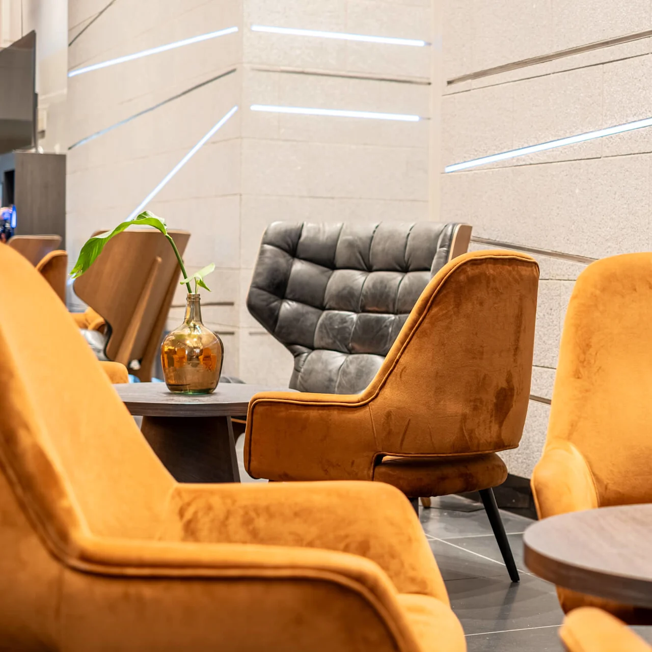 Cozy armchairs in various colors surround small round tables with glass vases holding green leaves, set against a modern, light gray tiled wall with subtle lighting accents.