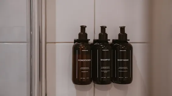 Tre mørke pumpeflasker mærket BALSAM, SHAMPOO og BODY WASH monteret på en hvid flisebelagt badeværelsesvæg.