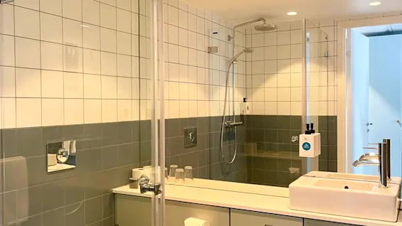 Et moderne bad med veggmontert toalett, vask og dusjområde. Rommet er innelukket med glasspaneler, flislagte vegger, og har stilige armaturer som skaper en ren, minimalistisk stil.