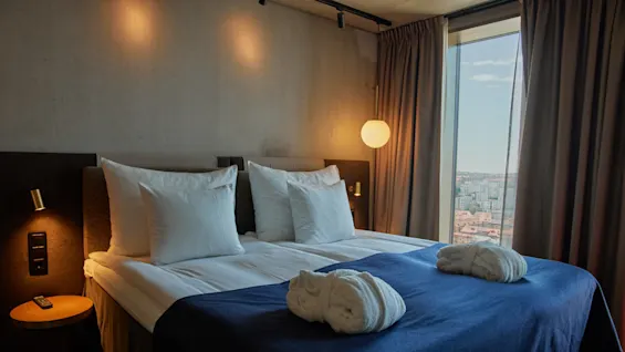 En prydligt bäddad säng med vita kuddar och en mörkblå filt har två vikta badrockar. Varm belysning och ett stort fönster med utsikt över staden kompletterar det mysiga hotellrummet.