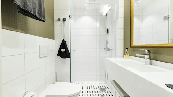 A modern bathroom features a wall-mounted toilet, a white sink with a faucet, gold-framed mirror, and a glass-enclosed shower. Black and white checkered floor tiles add contrast.