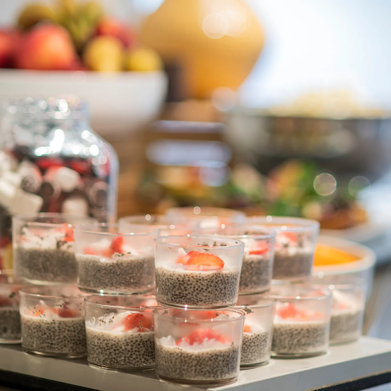 Chiapuddingskoppar toppade med jordgubbar är arrangerade på ett bord i en bufféinställning, omgivna av burkar och en skål med frukt.