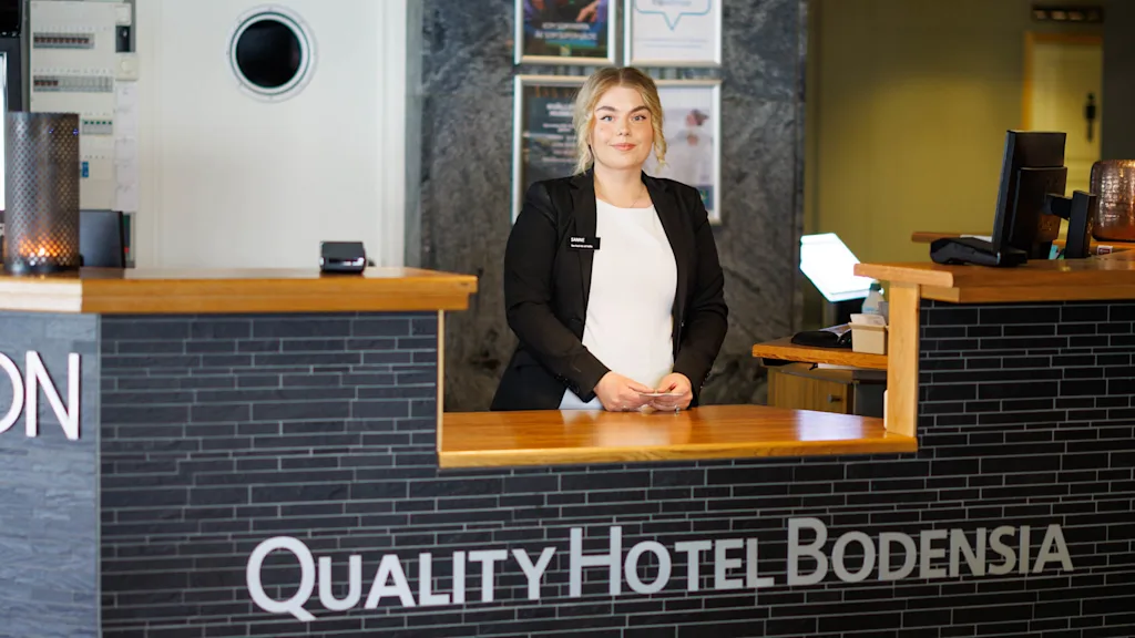 Vastaanottovirkailija seisoo hymyillen hotellin vastaanottotiskin takana modernissa aulatilassa. Tiskin kyltti lukee: “QUALITY HOTEL BODENSIA.” Taustalla on seinäkello ja mainosjulisteita.