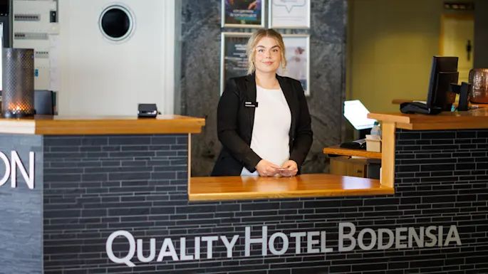 En resepsjonist står smilende bak hotellskranken i en moderne lobby. Skiltet på disken lyder: “QUALITY HOTEL BODENSIA.” En veggklokke og reklameplakater er synlige i bakgrunnen.
