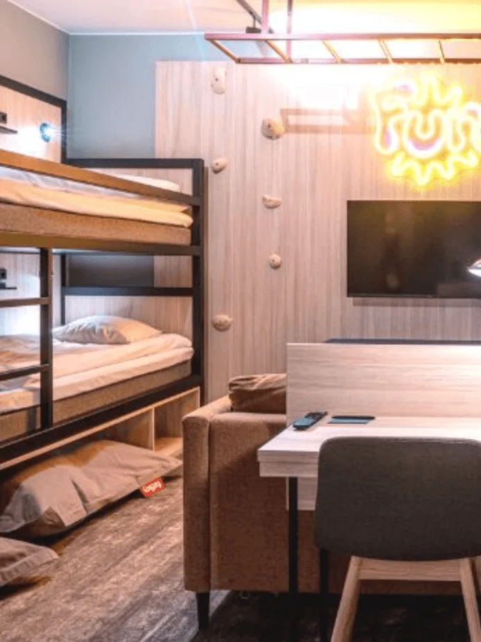 Våningssängar mot en vägg med klättergrepp. En neonskylt lyder "Fun". Ett närliggande skrivbord, stol och TV skapar en mysig rumsmiljö.