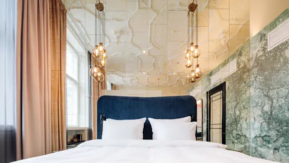 Bed with white linens, standing beneath hanging glass light fixtures. Mirrors and marbled walls surround the cozy modern room, with large curtains framing a window.