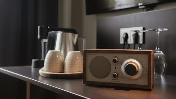 Radio står på et mørkt bord, omgivet af to kopper på en bakke, en kaffemaskine og et opadvendt glas i et moderne hotelværelse med blød belysning.