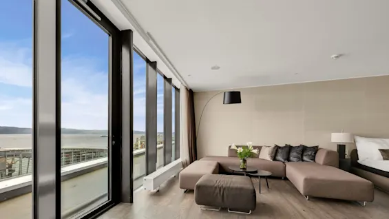 A large corner sofa faces a modern living room with floor-to-ceiling windows overlooking a waterfront. A small table with a vase of flowers sits in front.