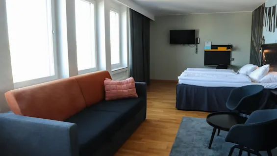 En mjuk orange soffa med en rosa kudde står i ett modernt hotellrum. I närheten finns en säng och en stol på trägolv under ljusa fönster.