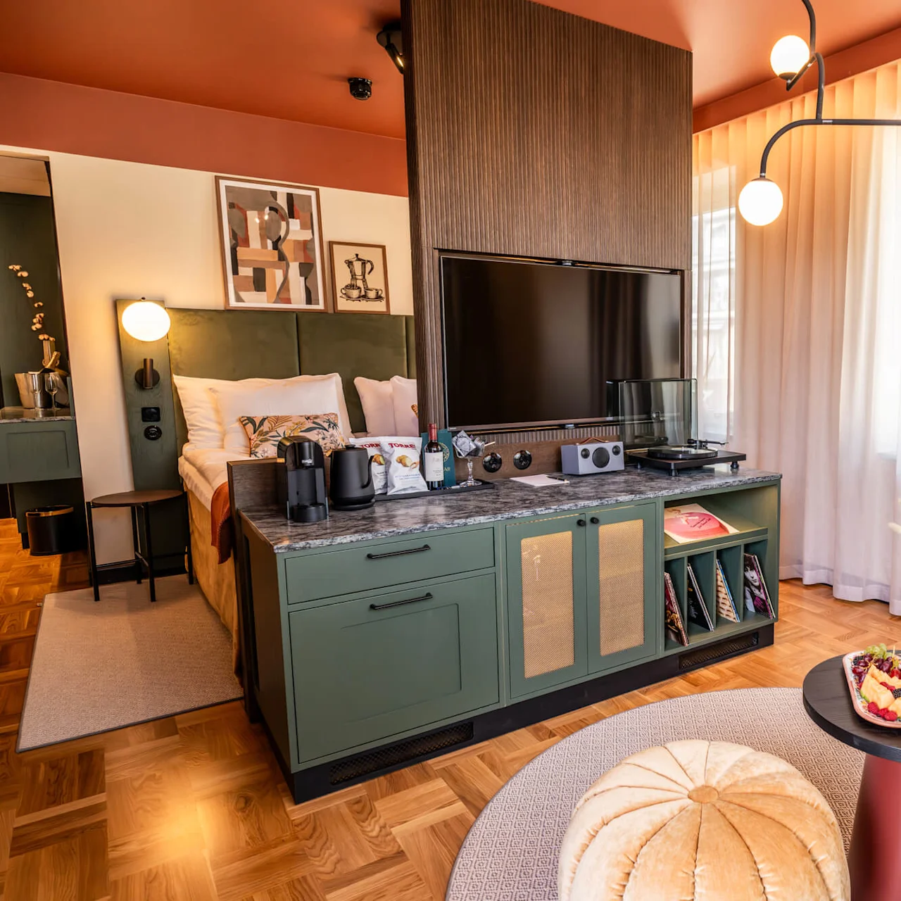 A cozy hotel room with a neatly arranged bed, large TV on a cabinet, armchair, and a table with fruit. Ambient lighting and modern decor create a warm, inviting atmosphere.