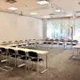Conference room with U-shaped arrangement of tables and chairs, surrounded by white walls and windows with curtains allowing natural light. Ceiling lights and a projector are visible.