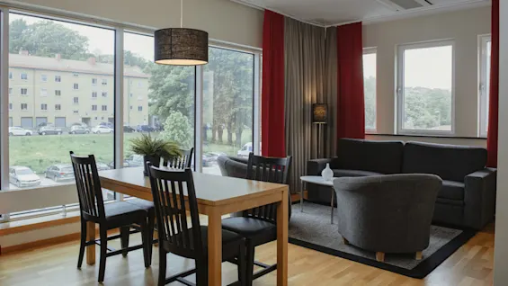 Dining table and chairs sit near large windows, providing a view of a green area with trees and apartment buildings. A cozy seating area with a sofa is adjacent.
