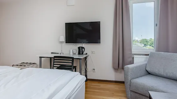 Seinäkiinnitteinen TV pöydän yläpuolella, jossa on lamppu ja vedenkeitin. Sijainti on hotellihuone, jossa on sänky ja sohva ikkunan vieressä, josta näkyy ulkoilmanäkymä.