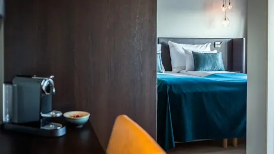 Seng med teal-farget teppe og puter, mykt opplyst av hengelamper, ved siden av et trebord med kaffemaskin og en skål; moderne soveromsmiljø.