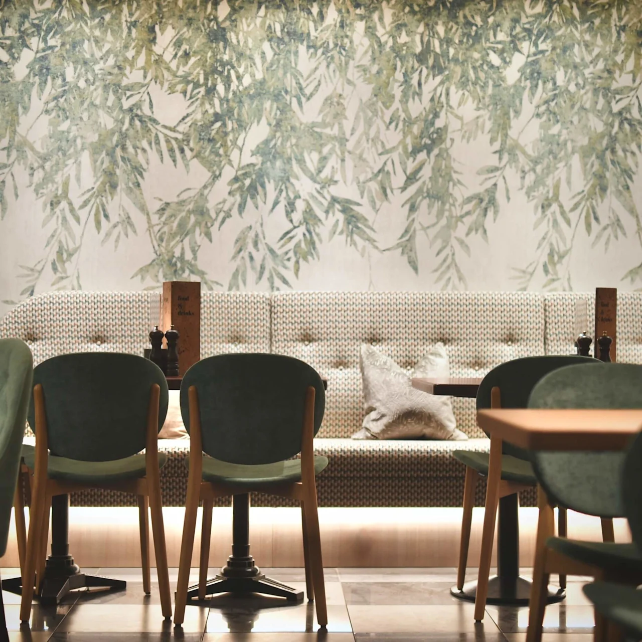 Gröna vadderade stolar är placerade runt bord i en mysig kafémiljö med lövigt tapet och mjuk belysning. Menykortet lyder "Food and Drinks".