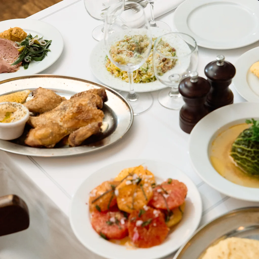 Et dækket spisebord med hvid dug fyldt med forskellige retter, herunder stegt kylling, salater og andre tilberedte retter, klar til et måltid.