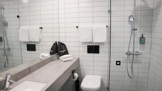 Et moderne baderom har servant, dusj og toalett. Hvite fliser dekker veggene, med hengende håndklær. Vesker merket “HAIRDRYER” og “LAUNDRY” ligger på benken.