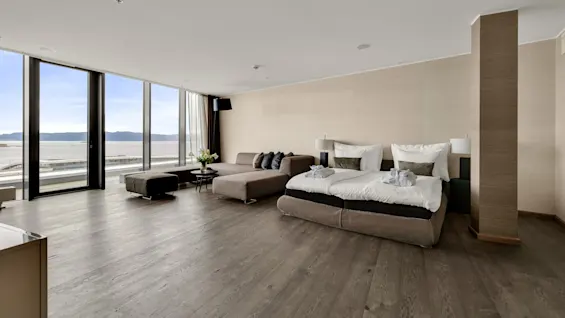 Luxurious bedroom features a large bed with pillows and robes, complemented by a chaise lounge. Floor-to-ceiling windows provide a sea view, enhancing the spacious, minimalist decor.