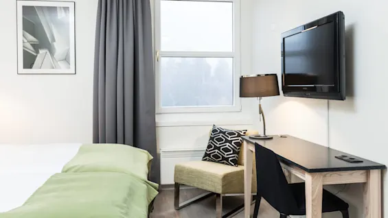 A neatly arranged hotel room features a neatly made bed with green covers, a desk with a table lamp and chair, a wall-mounted TV, and a window with grey curtains.
