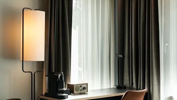 Et koselig hotellromhjørne med et skrivebord, kaffemaskin, radio og lamper, badet i naturlig lys fra et vindu med gardiner.
