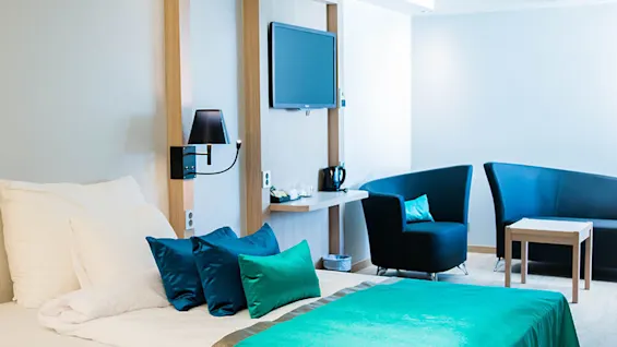 Seng med turkis og blå puder står nær vægmonteret TV; tilstødende sort stol og bord fuldender moderne hotelværelsemiljø.