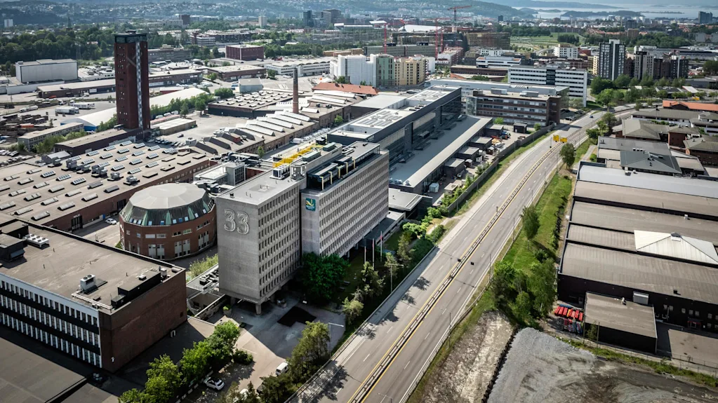 Bygning med "33" på står midt i et vidstrakt industriområde, omgitt av veier og andre store bygninger, under en delvis skyet himmel.