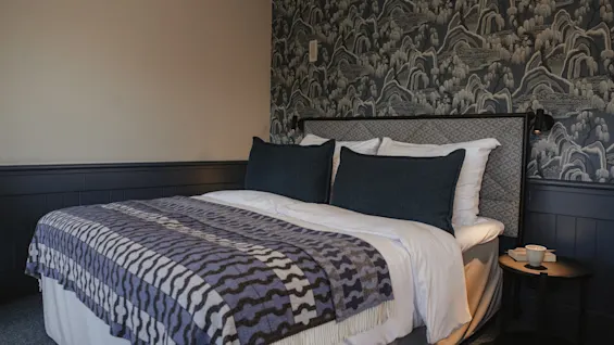 Ett mysigt sovrum med en säng med en mönstrad filt, mörka kuddar och en unik tapetdesign bakom sänggaveln.
