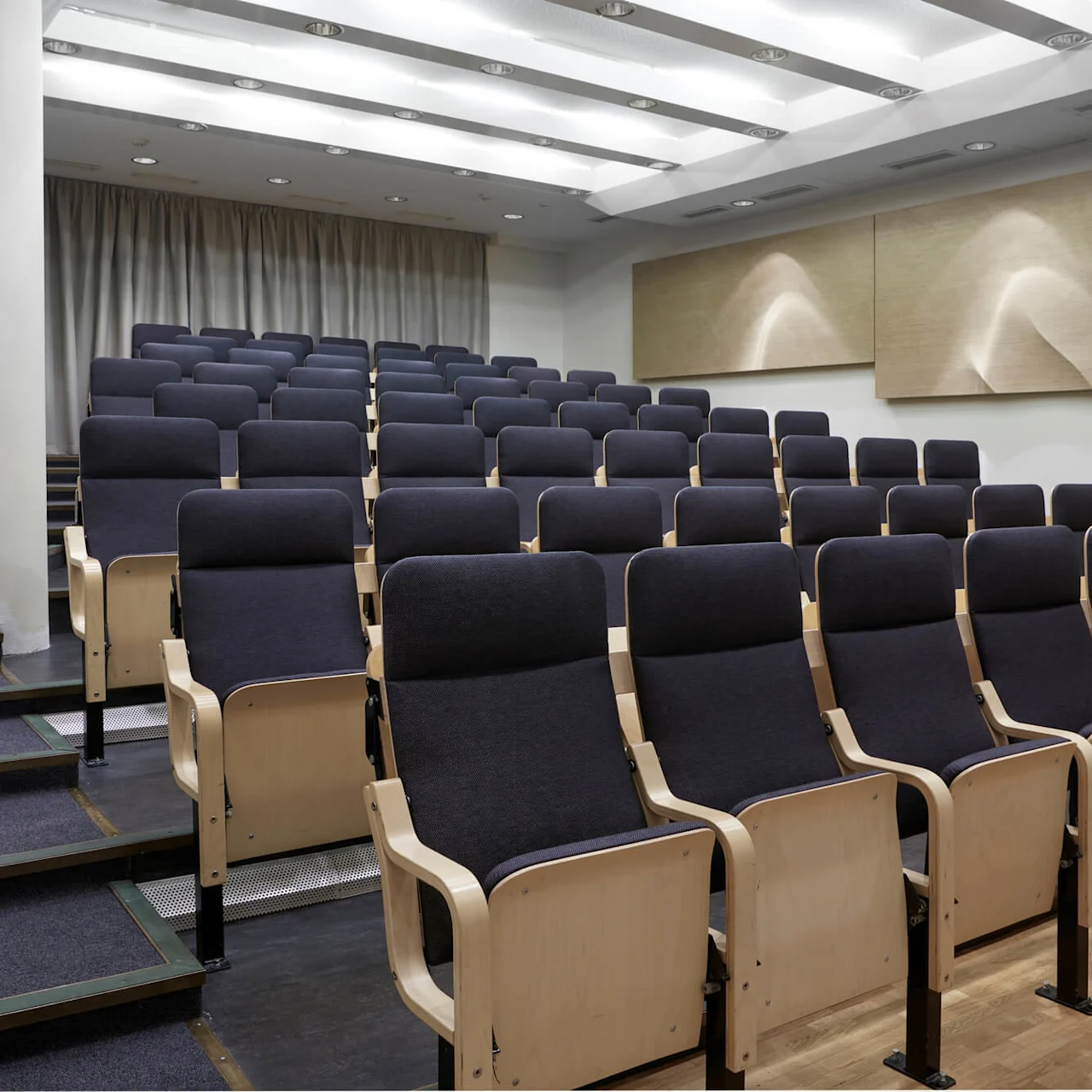 Rows of empty, dark-upholstered lecture hall seats are arranged on tiered flooring, surrounded by white walls with acoustic panels. The ceiling features modern lighting, providing bright illumination.