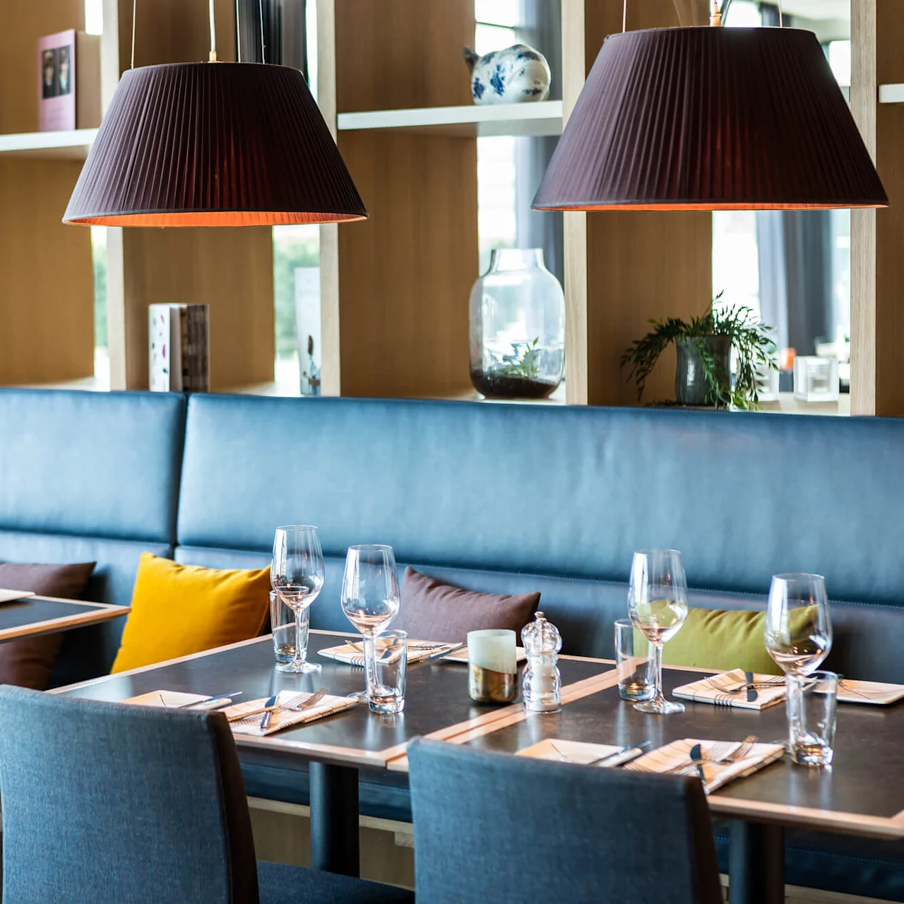 Tables are set elegantly with glasses and napkins, surrounded by upholstered seating. Shelves with decor and large pendant lights hover above. Cozy, modern restaurant atmosphere.