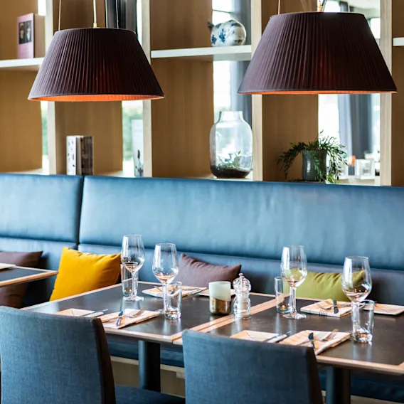 Bordene er elegant dekket med glass og servietter, omgitt av polstrede sitteplasser. Hyller med dekorasjoner og store pendellamper over. Koselig, moderne restaurantatmosfære.