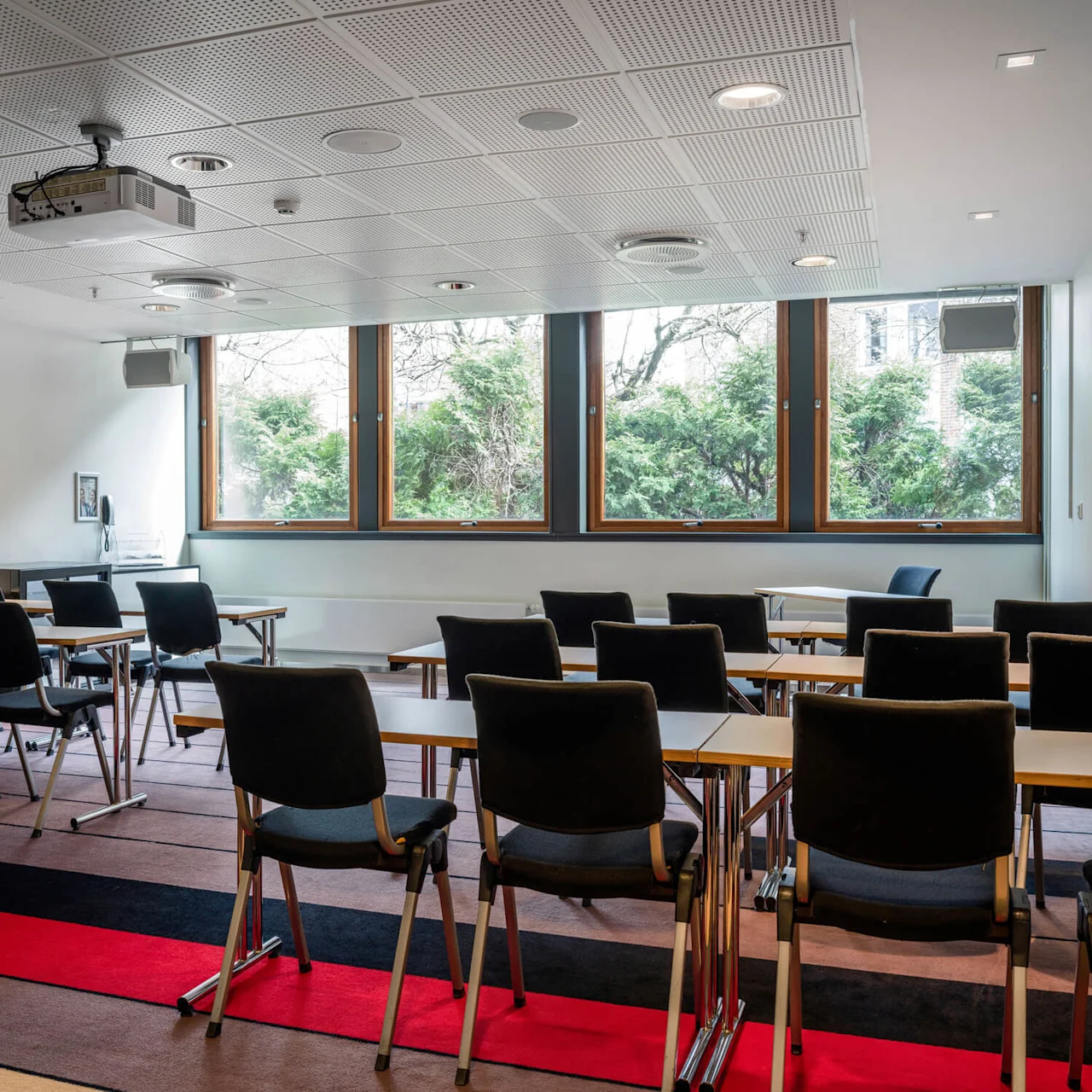 Classroom with empty chairs and desks facing a ceiling-mounted projector. Large windows reveal greenery outside, and the floor has a striped red and black pattern.