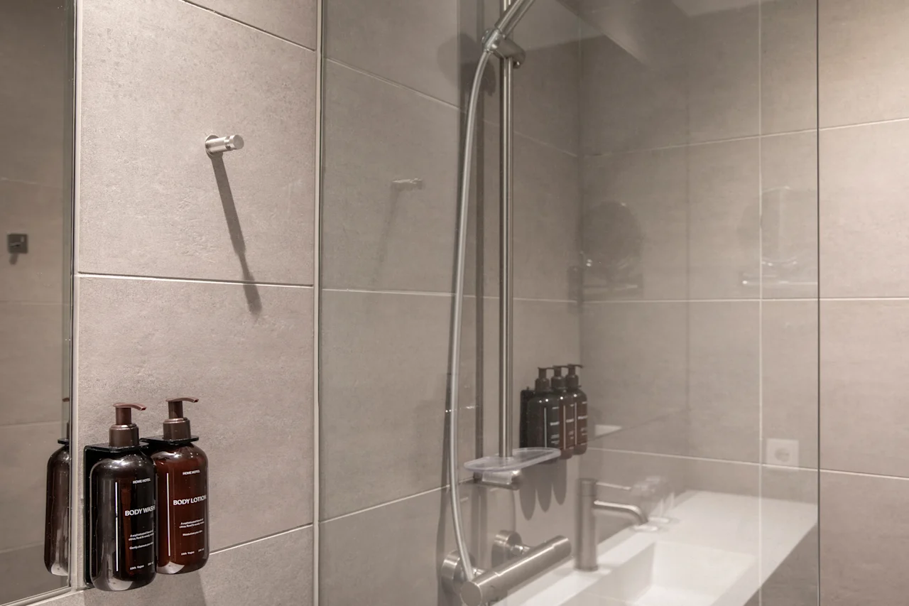 Et moderne bad med grå fliser, en dusj og en vask. To pumpeflasker merket BODY WASH og BODY LOTION er montert på veggen.