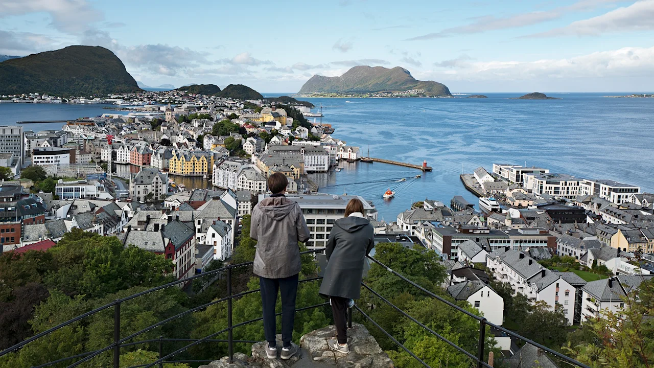 Two people stand on a hill overlooking a coastal city with colorful buildings, surrounded by water and distant mountains under a partly cloudy sky.