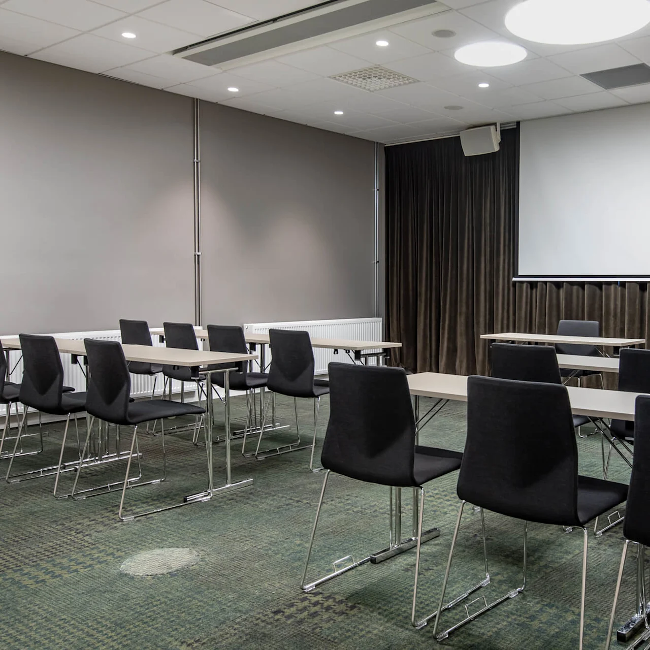 Rows of empty black chairs face a blank projection screen; tables are arranged in a conference room with gray walls and a green carpet. Ceiling lights illuminate the space.
