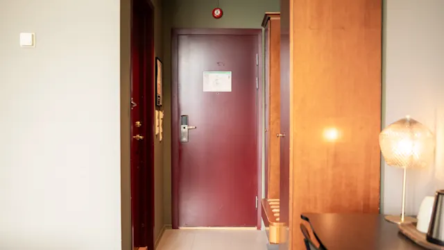 En lukket rød dør med et håndtak i et hotellrom, omgitt av tremøbler og en lampe på et skrivebord. Et lite skilt er synlig på døren.