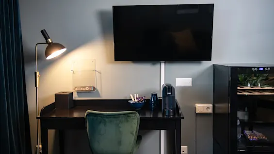 A desk with a green chair holds a coffee machine and bowl, beside a lamp. A TV is mounted above, and a fridge is nearby in a cozy room.