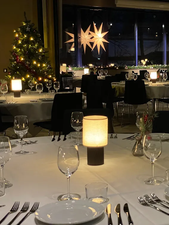 Elegant tables are set with white cloths, plates, and glassware. Soft lighting and illuminated decorations create a festive atmosphere in a spacious dining room.