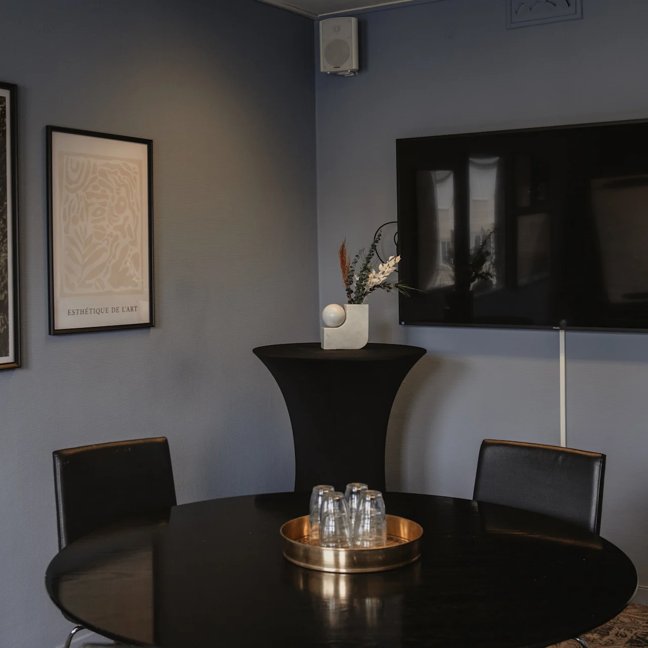 A round black table with a tray of glasses stands in a room with blue walls. Two framed pictures, a large TV, and a decorative pedestal are nearby.