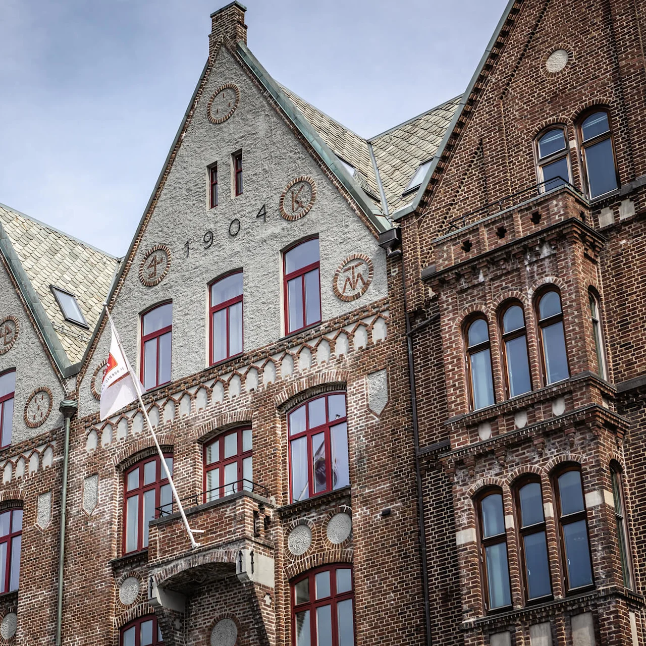En historisk mursten- og stenbygning med rødrammende vinduer i Bergen. Teksten "BRATTEN" og "1904" er synlig. Et flag hænger fra en stang på facaden under en klar himmel.