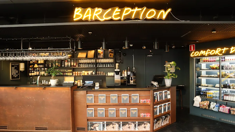 A bar with illuminated "BARCEPTION" sign above and a "COMFORT B" sign on the right. Magazine racks and a display fridge with snacks are part of the setting.