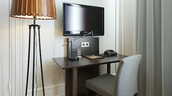 A well-lit modern desk with a television, lamp, phone, and notepad, set against a patterned wallpaper and beside a window with curtains.
