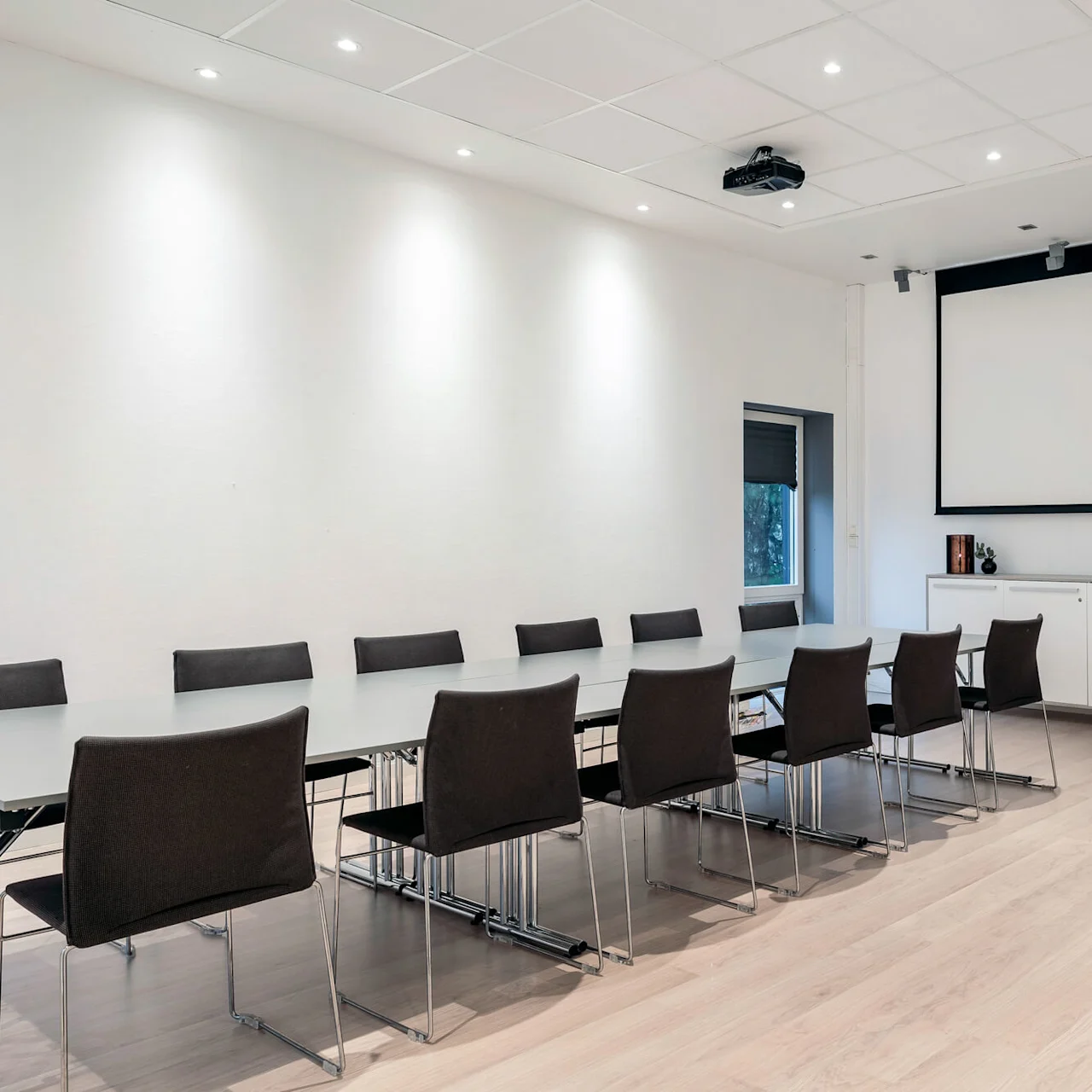 Et langt rektangulært konferansebord er omgitt av sorte stoler, vendt mot en stor hvit projeksjonsskjerm. Rommet er moderne med tregulv og minimal innredning.