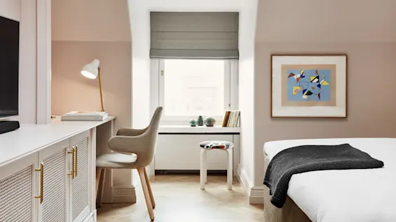 Et skrivebord med lampe lyser opp bøker ved siden av en stol under et vindu med vaser og bøker; soverom med abstrakt kunst på veggen. Tekst: "THE DESIGN HOTELS™ BOOK".