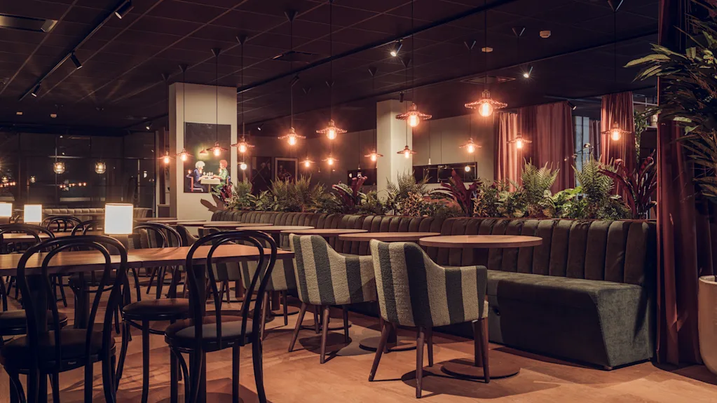 En restaurang har träbord och stolar arrangerade snyggt, omgiven av mjuk belysning. Krukväxter pryder utrymmet och skapar en mysig stämning med varma toner och modern inredning.
